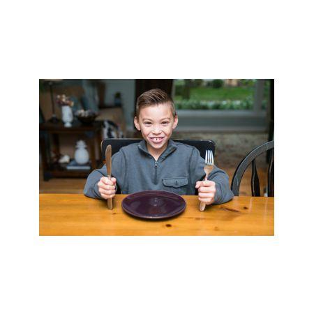 Smiling child with cutlery sitting at a wooden table, empty plate in front